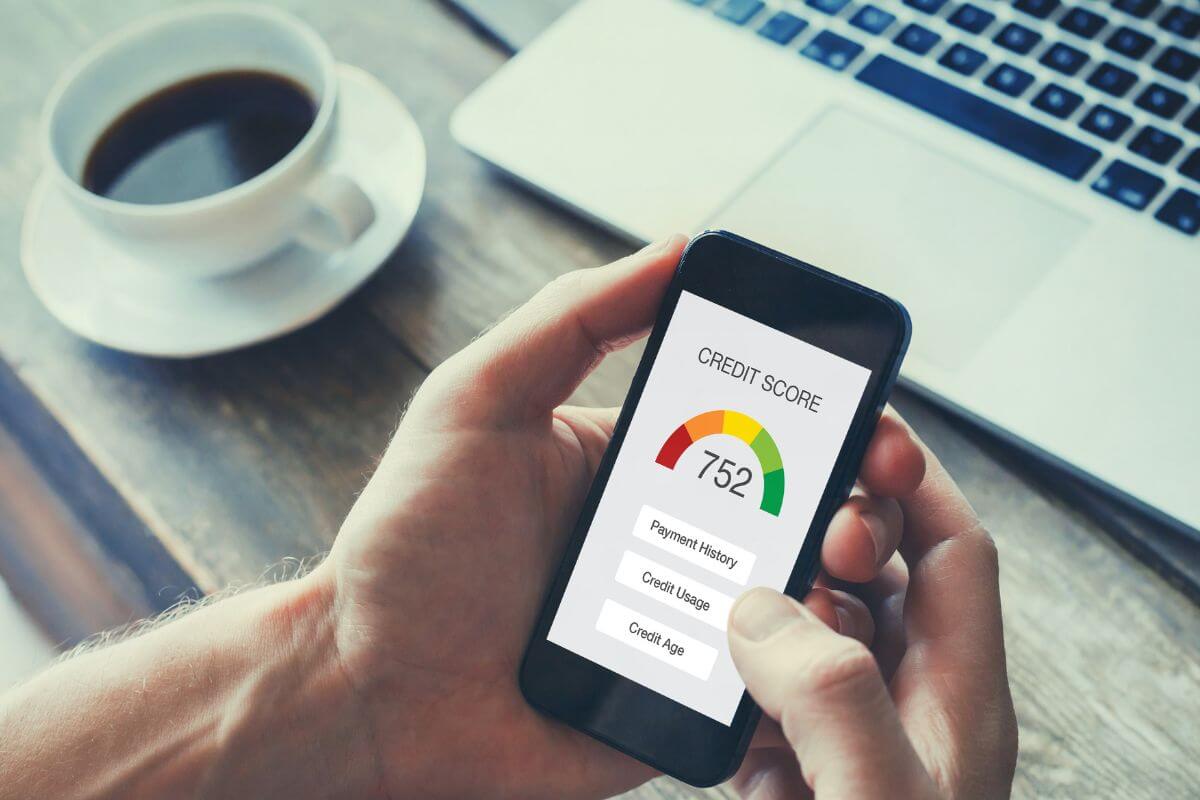 The image shows a coffee on the table, a laptop, and a man's hand holding a phone with a screen display which means to build or improve your credit score saves you money on loans and credit cards.