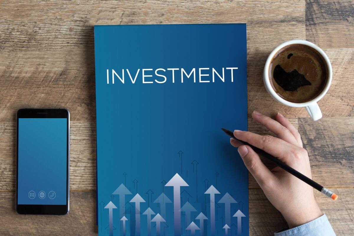 The picture tells about a coffee, a phone, a book with a title, and a man's hand holding a pen indicating that investments are diversified and aligned with your goals that ensure you're on track for long-term success.