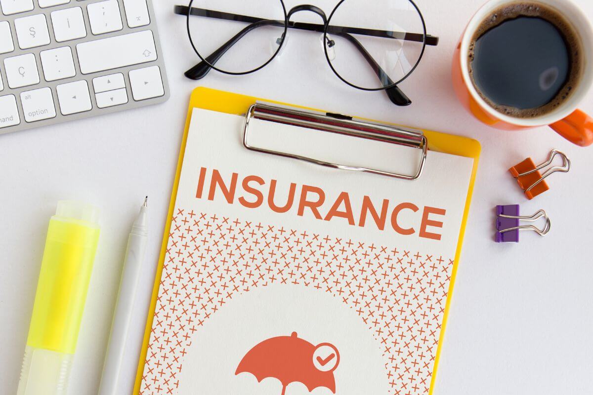 The image shows a keyboard, a coffee, a marker, a pen, clips, an eyeglass, and a clipboard with notes referring to having adequate insurance coverage ensuring you're prepared for unexpected events and can cover without financial stress.