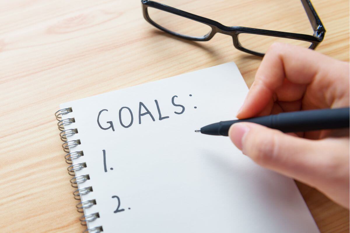 The picture of an eyeglass and a man's hand and writing on the notebook which means to set a financial goal keeps you motivated and easier to save when you know you're working toward and why it matters.