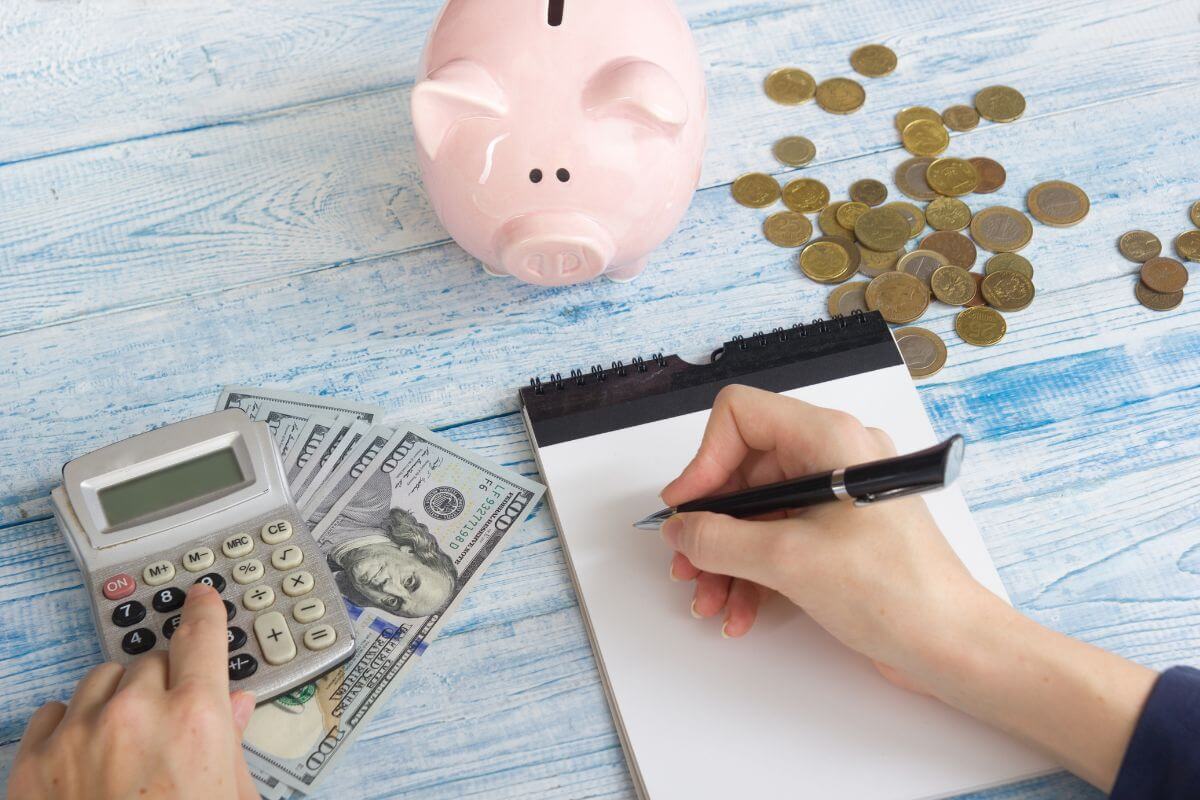 The picture of a piggy bank, coins, dollar bills, and a girl's hand using a calculator and writing on the notebook indicates to try the anti-budget by focusing only on saving a set amount and letting your spending take care of itself.