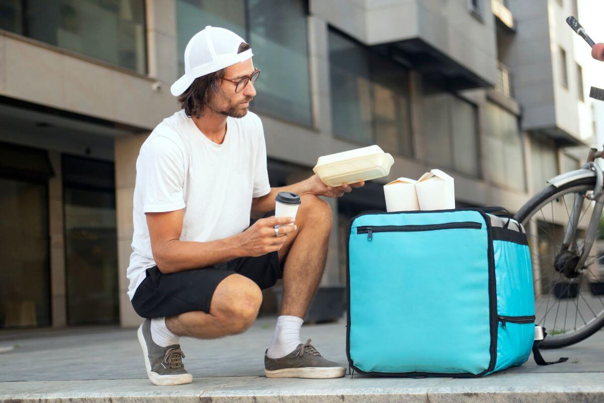 The picture shows a food bag container with food papers on the top and a man holding a food pack and coffee to deliver to his customer's address indicating to start a side hustle which gives you financial breathing room and a great way to work toward financial freedom.