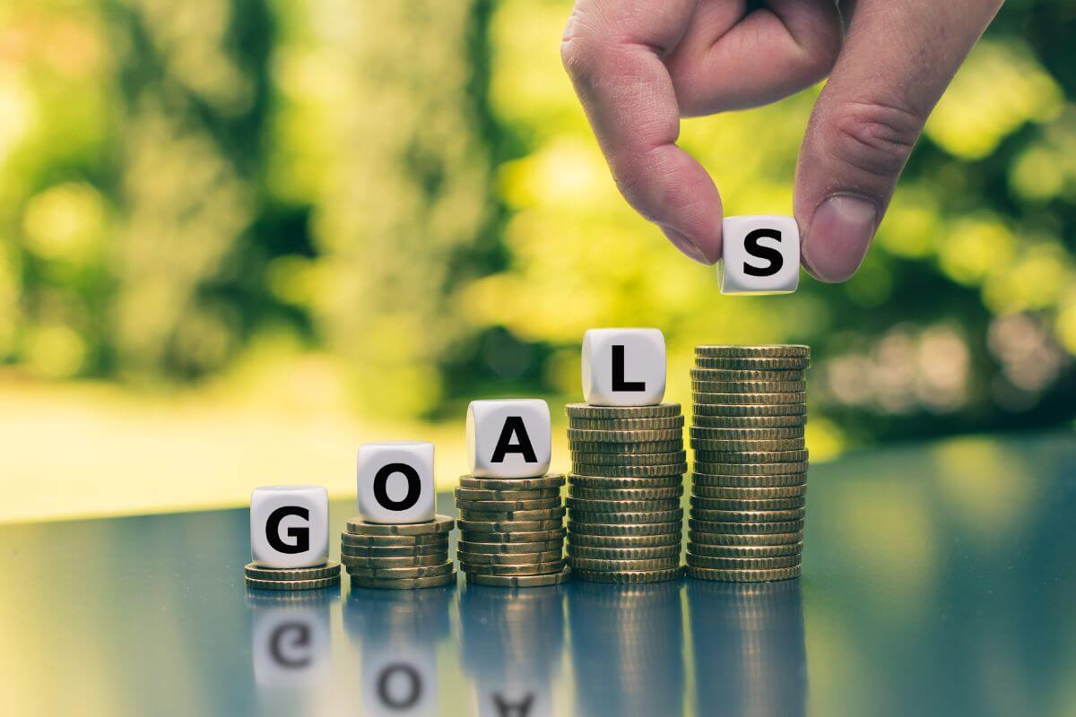 The image shows coin stocks, white cubes with letters, and a man's hand placing the letter s on the last stock coins which means to gamify your savings goals by setting small goals and rewarding yourself when you hit them.
