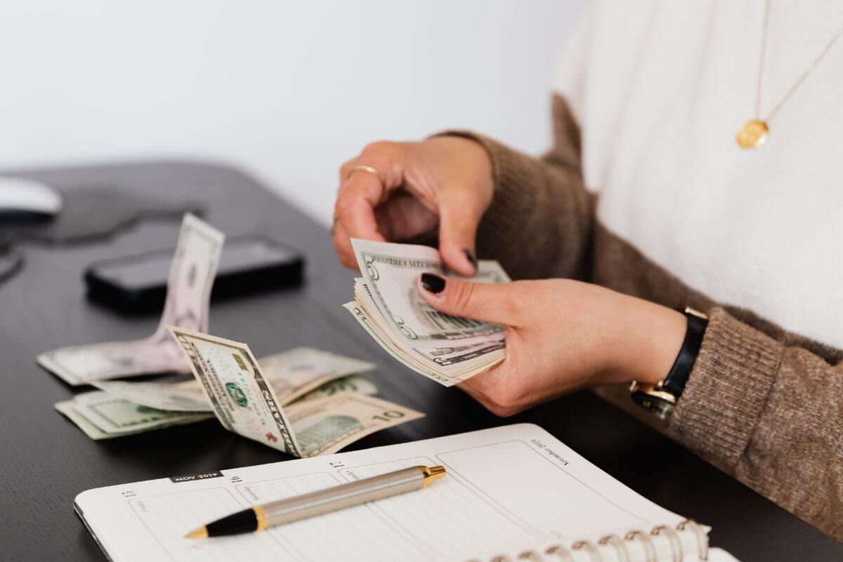 The photo tells about a phone, a notebook, a pen, and a girl counting and dividing dollar bills into groups. This means trying a zero-category budget by focusing on covering your fixed costs and savings, and then the rest of your spending flows naturally.