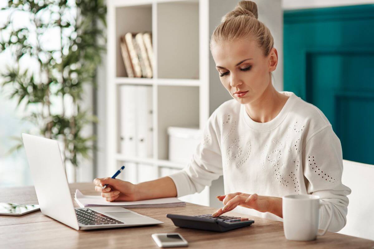 The photo shows a laptop, a phone, a cup, and a girl writing on the paper while calculating which means budgeting in reverse by analyzing past spending and adjusting forward to figure out what works for you without starting from scratch. 