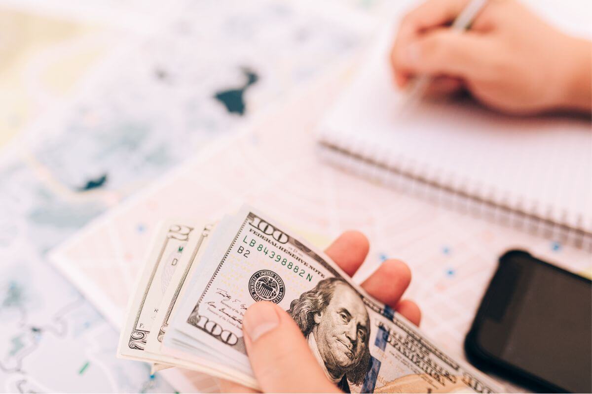 The image shows a phone and a girl's hand and writing on the notebook while holding dollar bills which means only budget by percentages of income so you know where every dollar goes without overthinking.