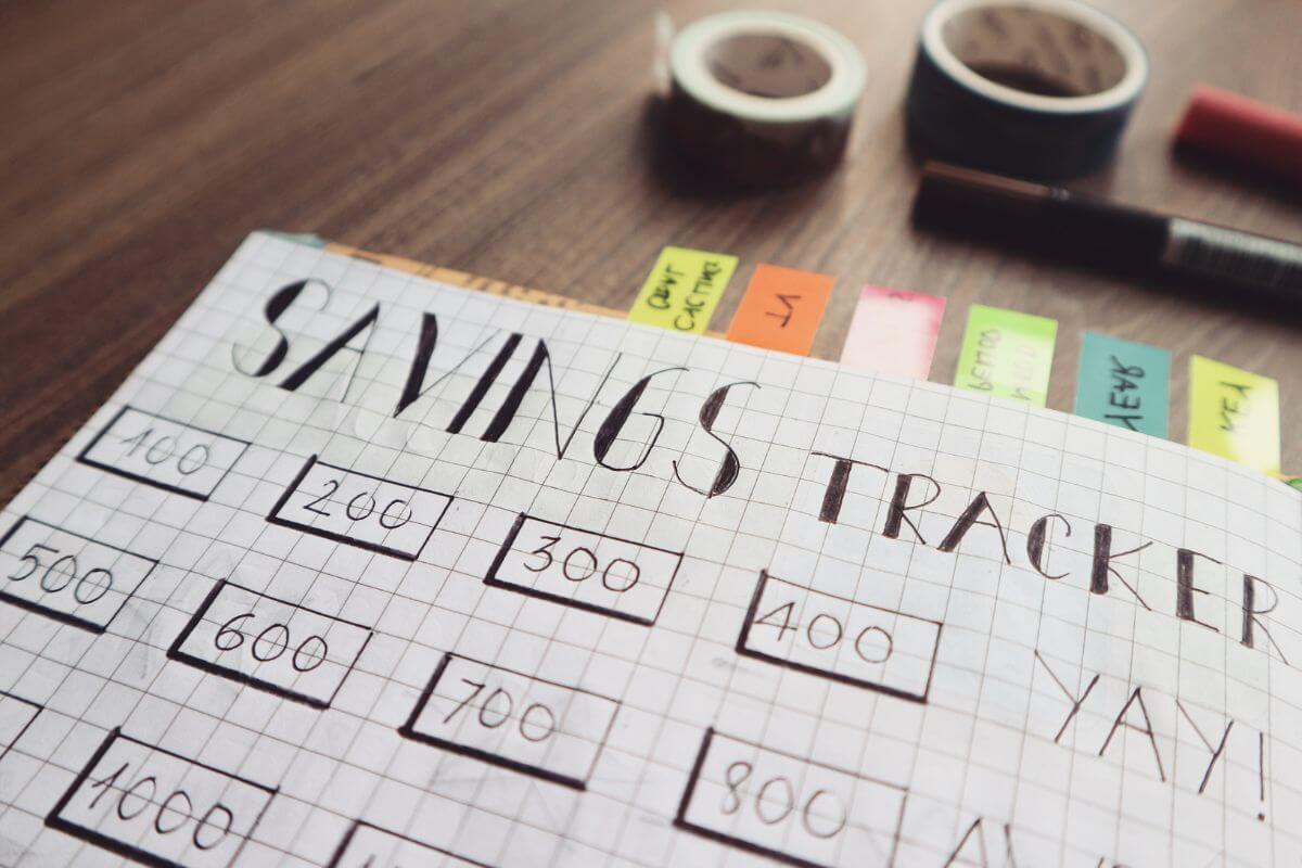 The photo shows tapes, pens, and a notebook with sticker notes and notes indicating to use visual tool like a saving chart helps keep you motivated and excited about reaching your goals.