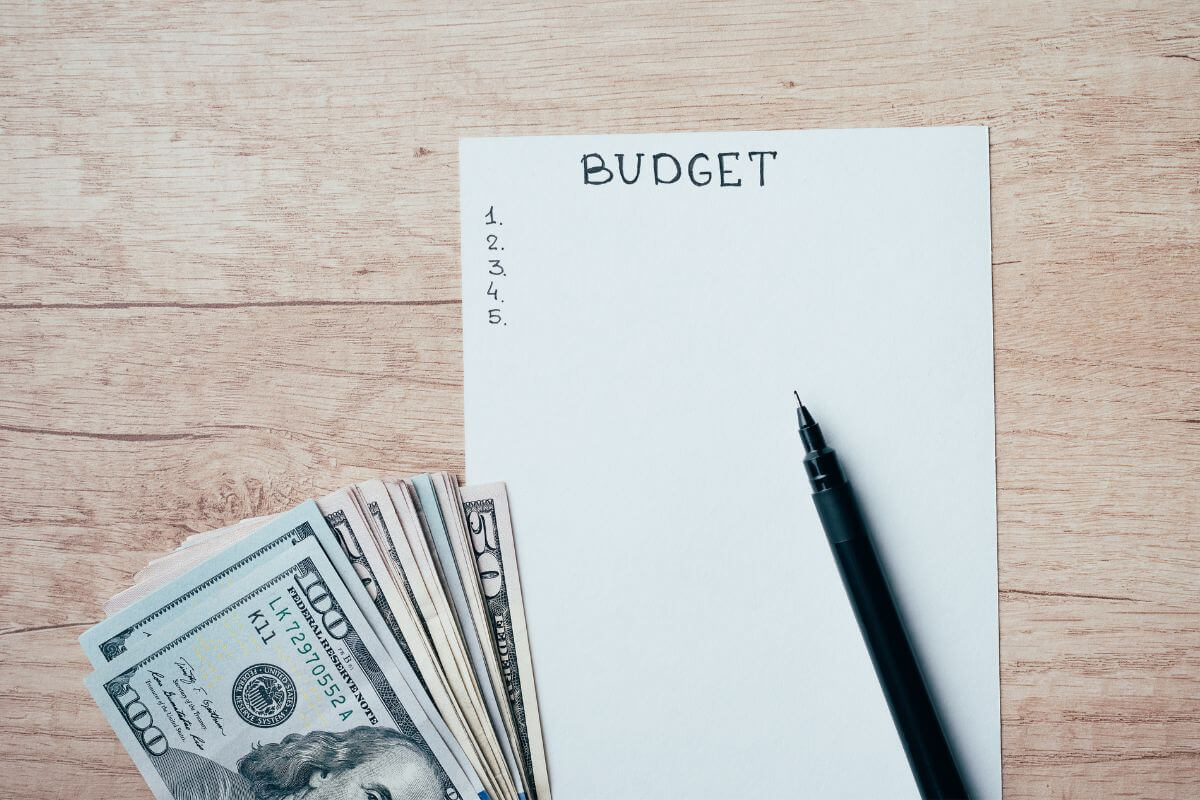 The image shows dollar bills, a pen, and a piece of paper with notes referring to treating budgeting like a game with challenges to keep yourself motivated and build better habits over time.