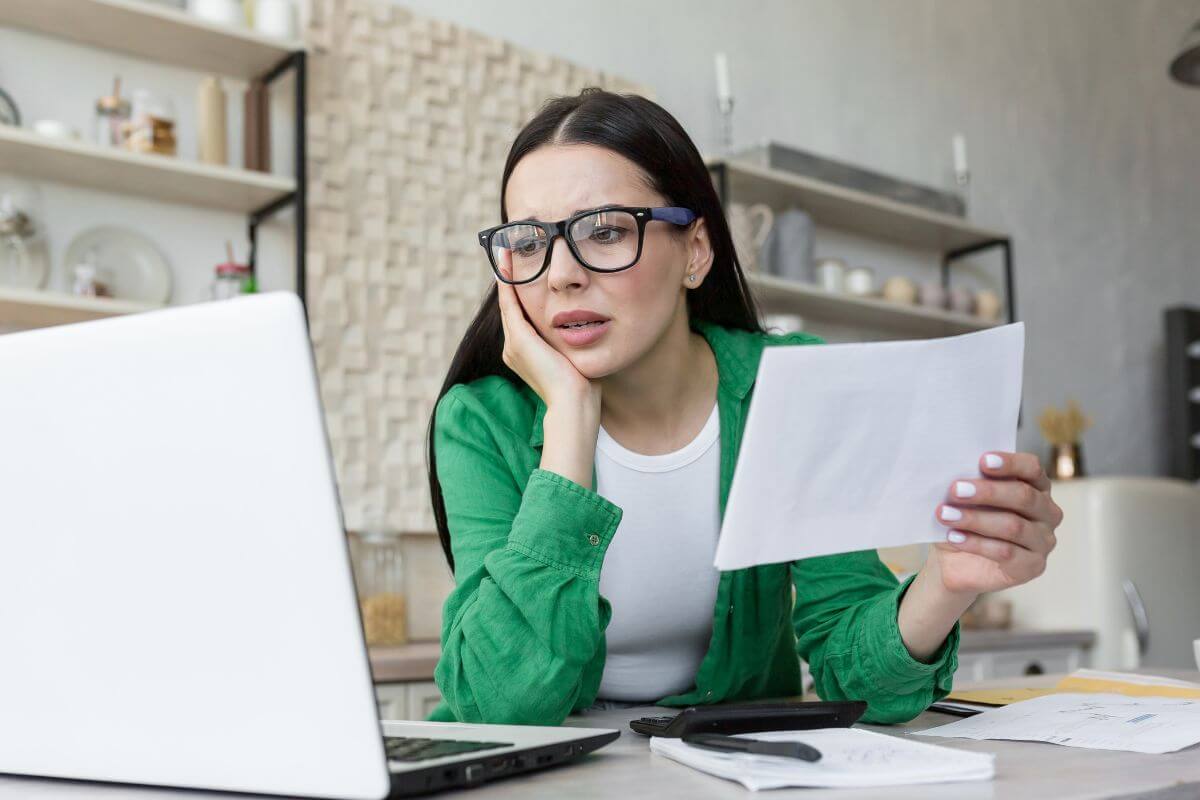 The image of papers on the table, a pen, and a girl holding a piece of paper while looking at the laptop means using the debt snowball method for creating quick progress and staying encouraged on your debt-free journey.