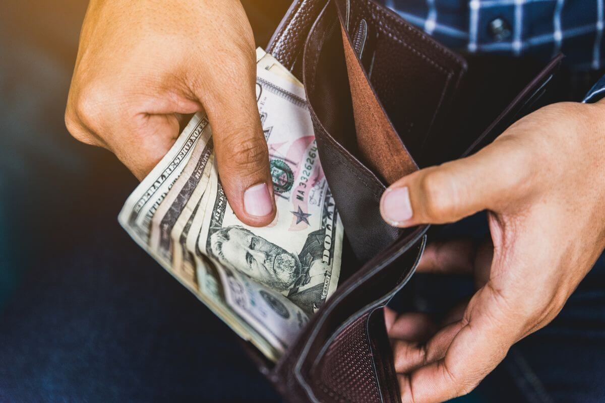 The photo of the man's hand pulling dollar bills on the wallet represents the implementation of a no-spend challenge for non-essentials for a set time and using extra cash to pay off debt.