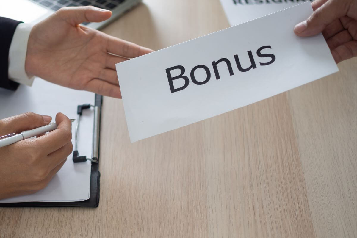The image tells about a man's hand handing the envelope with a note to a man's hand holding a pen with a clipboard indicating that dedicating all windfalls to debt payments helps reduce what you owe faster.