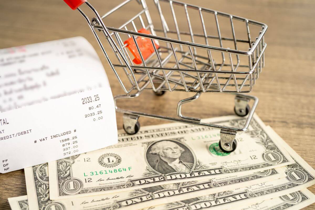 The image shows a receipt, a cart, and dollar bills which means to round up purchases and apply the difference toward your debt and an easy habit that can add up quickly without much effort.