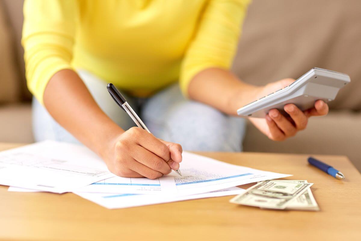 The image of dollar bills, a pen, and a girl's hand holding a calculator and writing on the paper which refers to paying biweekly instead of monthly can reduce interest and help you pay off debt faster.