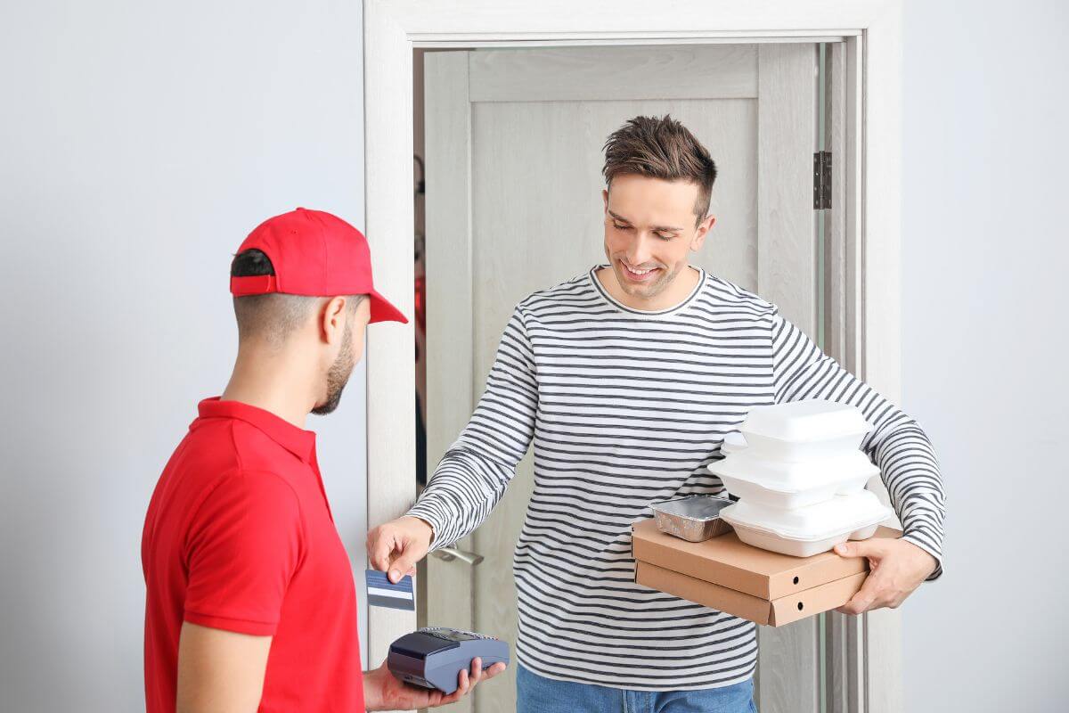 The picture tells about a customer holding his take out foods while tapping his credit card to a machine swipe held by a delivery man referring to allocate all expenses reductions toward debt to stay discipline and keep moving forward.