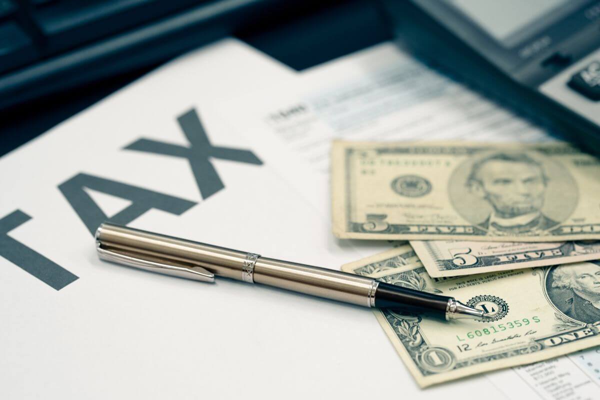 The picture tells about a calculator, dollar bills, a pen, and a piece of paper with a note indicating to use tax-loss harvesting that can offset gains and lower your taxable income.