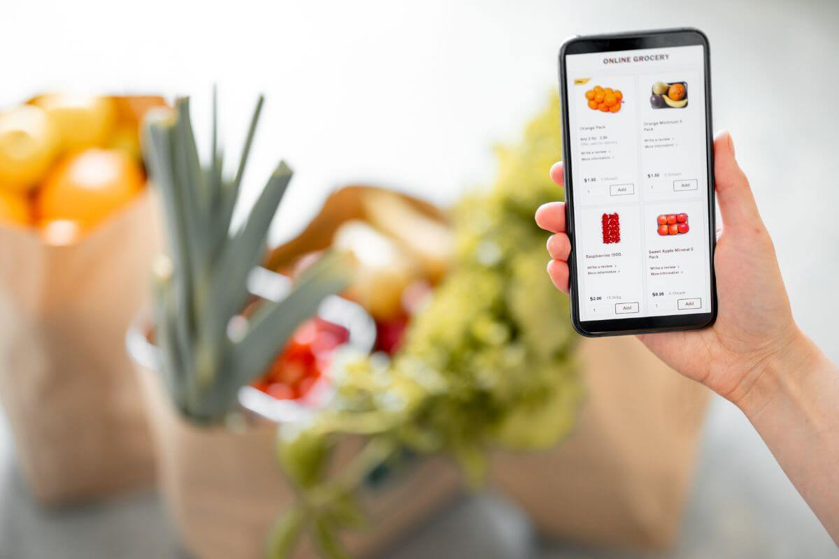 The photo shows fruits and vegetables behind, and a girl's hand using a phone with a screen display which means to plan grocery shopping around cashback or rewards apps to cut costs without sacrificing what you need.