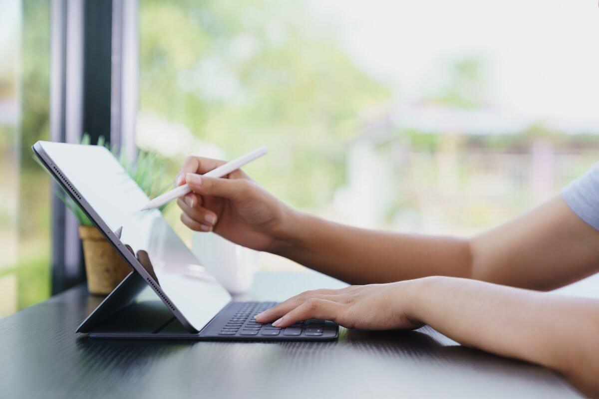 The photo shows a girl's hand using a digital tablet with a pen and keyboard referring to using subscription services for tools you need can save money and keep costs low.