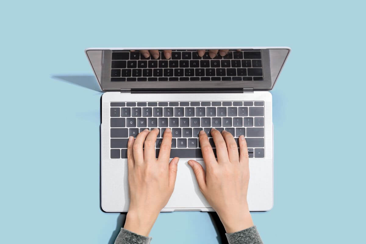 The image tells about a girl's hand using a laptop which means to create and sell digital products and a simple way to make money with skills you already have while working on your schedule.