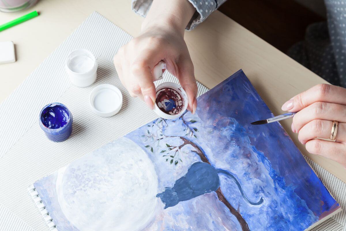 The image shows paints and a girl's hand painting the canvas with a cat themed which refers to offer personalized pet artwork that can grow your side gig while doing something you enjoy.