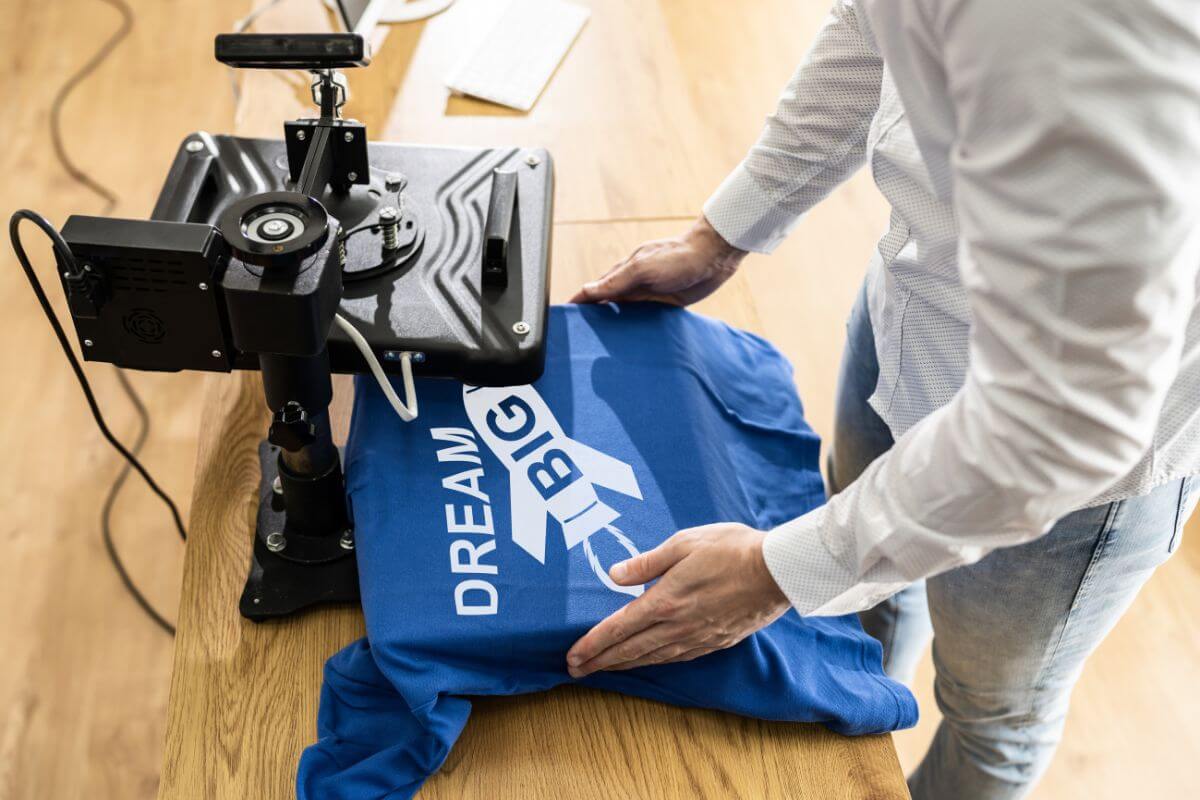 The photo of a man printing a short with the machine refers to designing and selling t-shirts or merchandise to start a side hustle with little upfront cost.