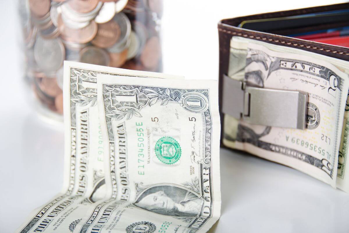 The picture of a jar filled with dollar bills, dollar bills on the table, and a wallet with dollar bills refers to setting a small daily spending limit and putting any leftover money toward debt.