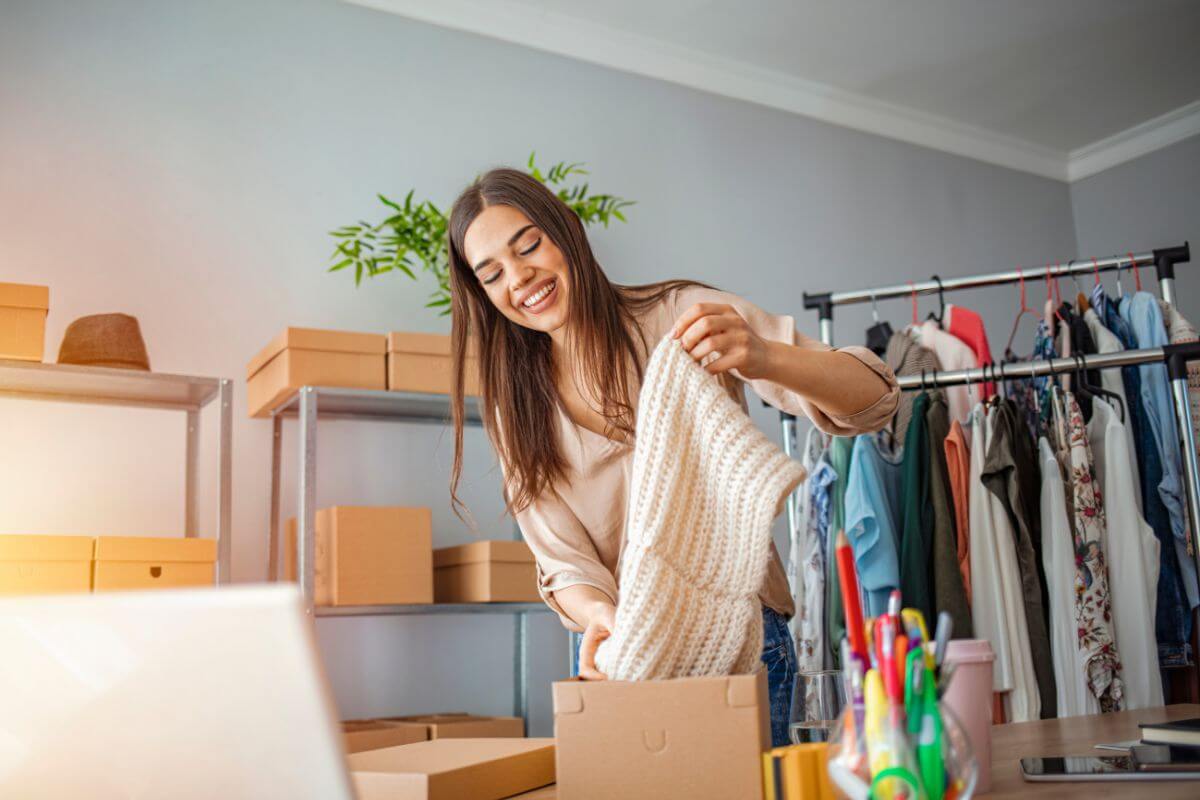 The photo shows boxes, clothes in the rack, pens, and a girl putting the clothes in a box which refers to flip thrifted or vintage items online, perfect to make money.