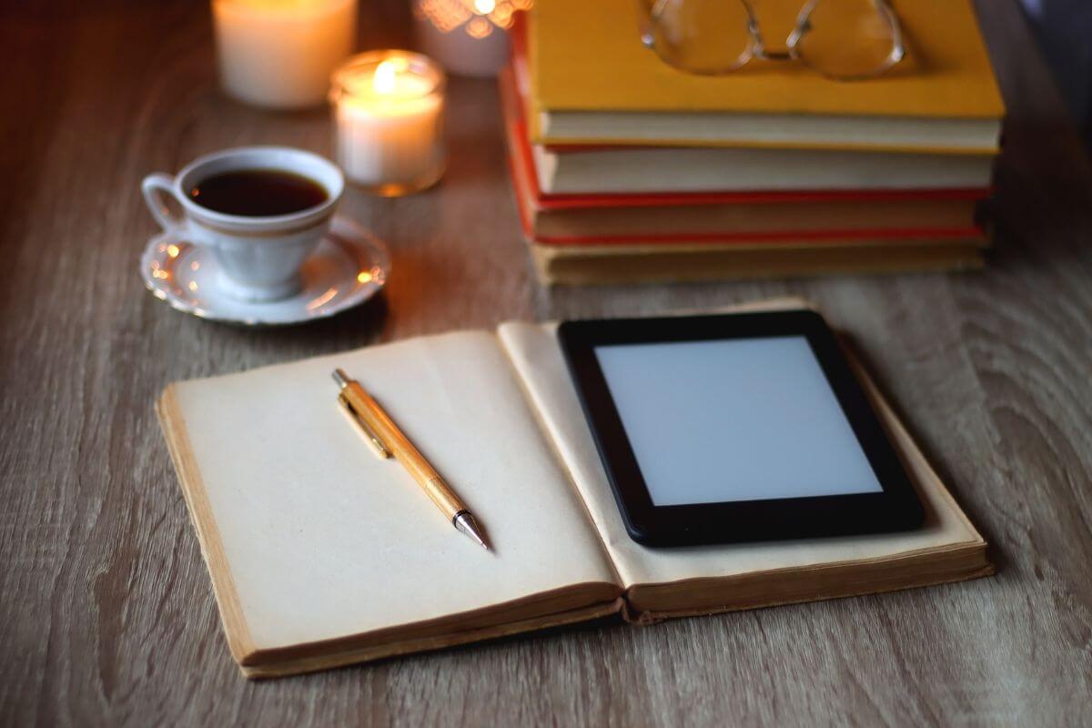 The picture tells about books, an eyeglass, candles, coffee, a pen, and a notebook with a tablet device indicating that writing and selling e-books is a great way to earn passive income while sharing valuable tips or insights. 