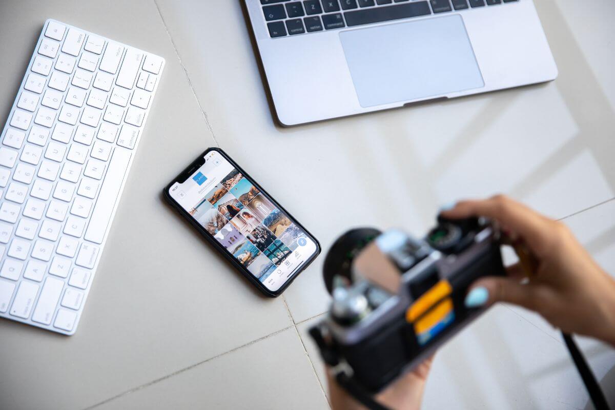 The picture of a laptop, a keyboard, a phone, and a girl's hand holding a camera indicates to create and sell of stock photos or videos and sell them online and make money.