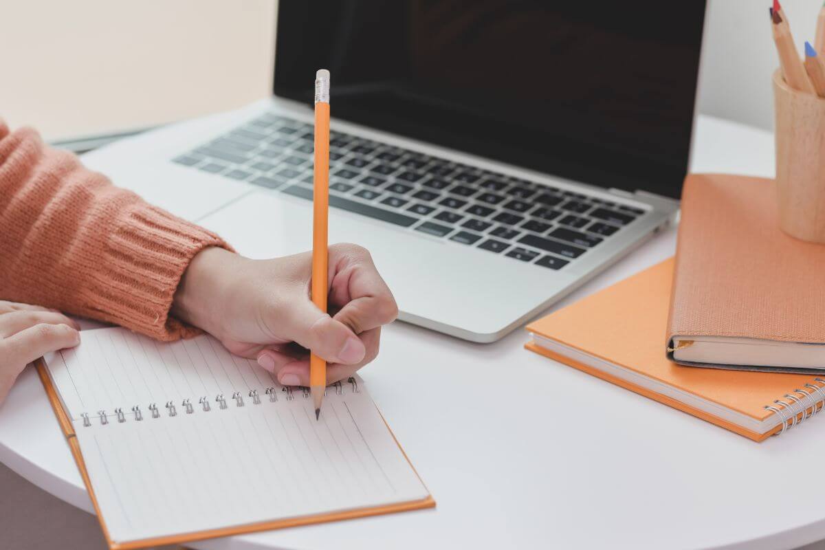 The picture of notebooks. A laptop is used for research, and a girl's hand is on a notebook which means to identify potential obstacles and plan solutions in advance that keeps you focused and avoid getting stuck.