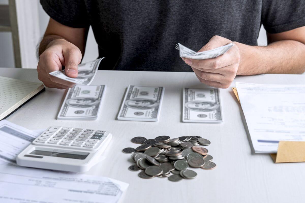 The photo shows papers, coins, a calculator, and a man dividing the dollar bills into groups which refers to a budget with a debt-first approach that helps you save on interest and reach financial stability faster.