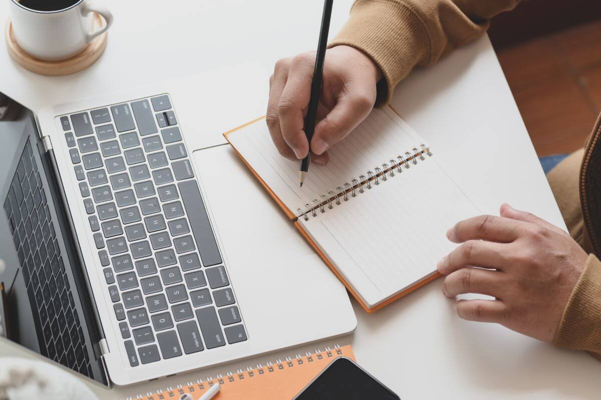 The image tells about a coffee and a man's hand that writing in a notebook while referring on the laptop refers to creating daily or weekly habits that align with your goals and make it easier to achieve what you set out to do.