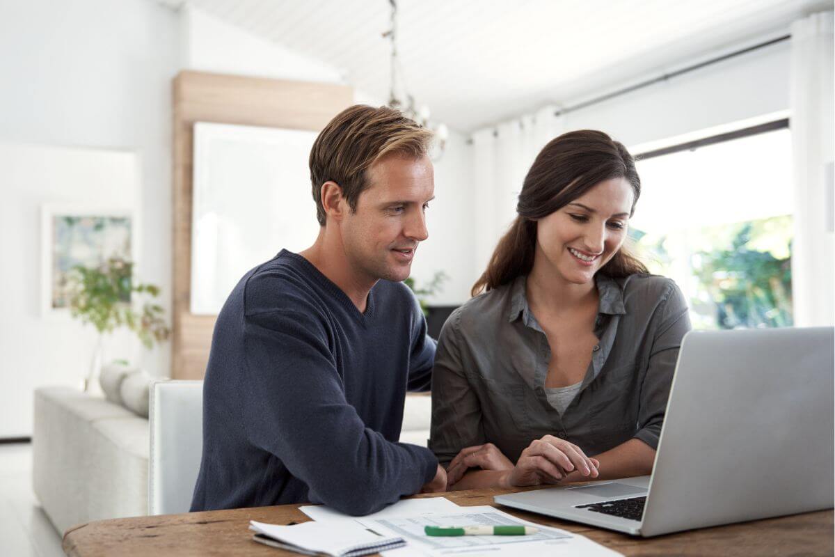 The photo shows papers on the table and a couple using a laptop together which means sharing your goals with a trusted partner can make the process less lonely and keep you on track.
