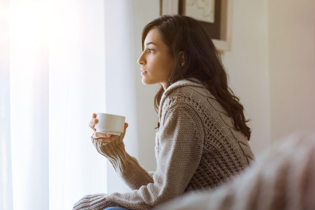 The photo of a girl holding a cap of coffee while looking at the window indicating to reflect on why the goal is important to you helps you stay connected to your purpose.