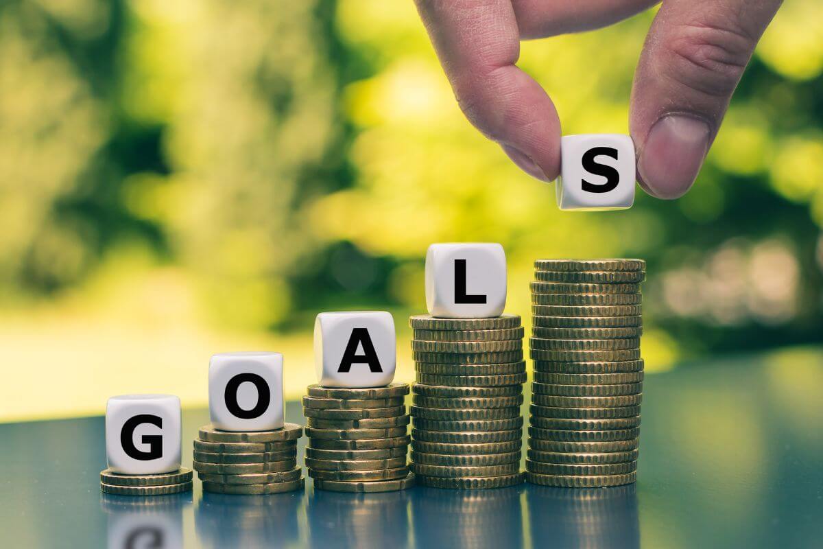 The photo tells about coin stocks, white cubes with letters, and a man's hand placing one on the last which refers to finding the best money goals can make real progress and set yourself up for financial success. 