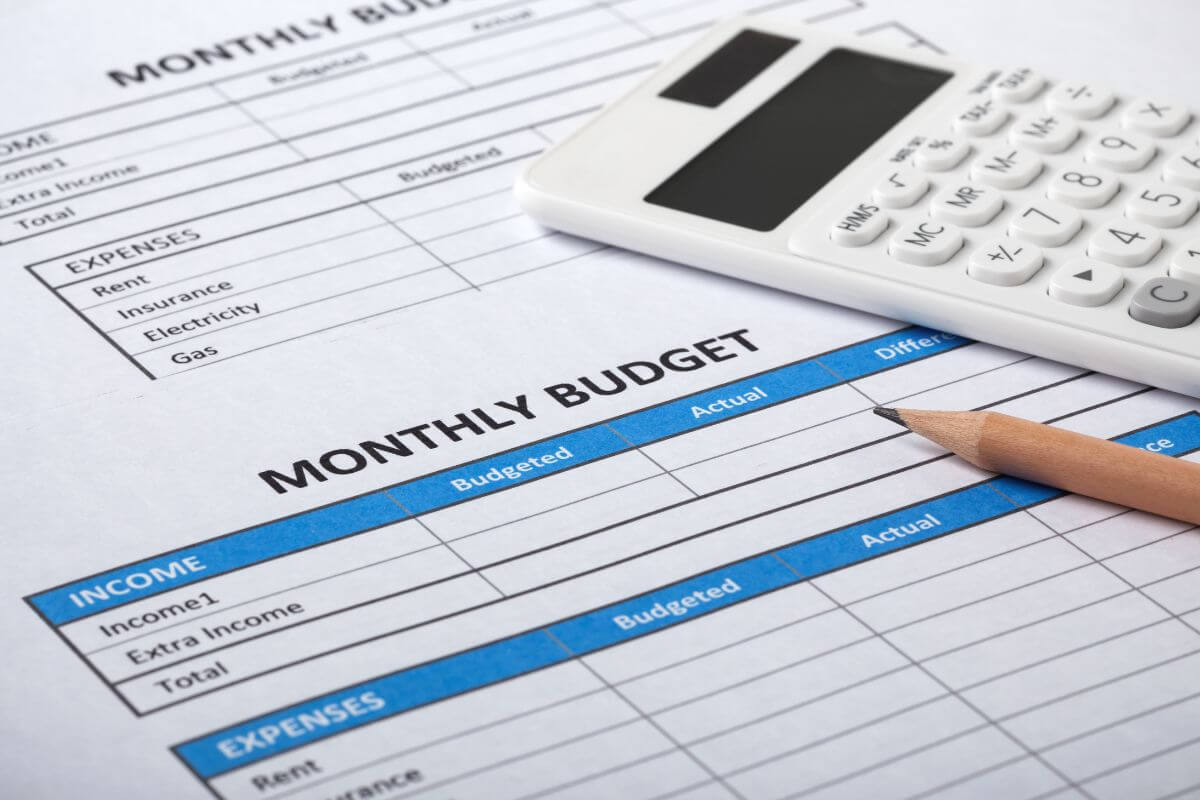 The photo shows a calculator, a pen, and papers with notes referring to creating and sticking to a monthly budget, the best ways to take control of your finances.