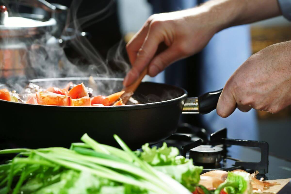 The picture shows vegetables, a kettle, and a man's hand cooking which means cooking meals at home instead of eating out which saves you money on expensive takeout while giving you full control over what you eat.