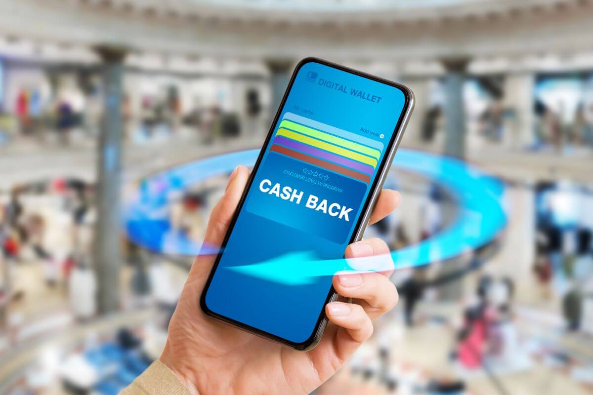 The image shows a girl's hand holding a phone with a screen display at the mall which means using cashback apps to make additional payments and apply them directly to your debt.