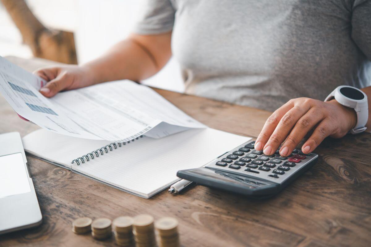 The picture shows coin stocks, a laptop, a notebook, a pen, and a girl's hand using a calculator while holding a paper indicating to track spending to identify unnecessary expenses and free up funds for saving or paying the debt.