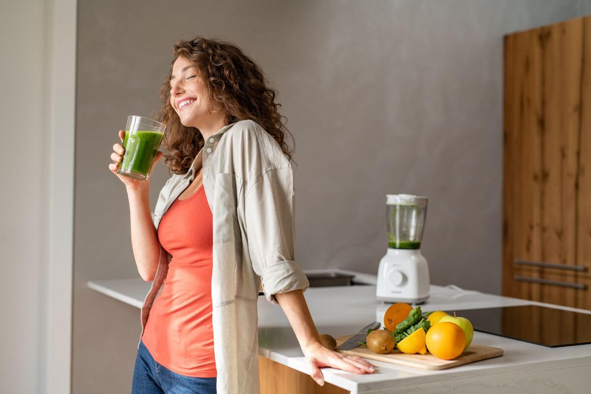 The photo shows fruits, a blender, and a girl holding a glass of drink which means making coffee and drinks at home which is a budget-friendly habit.