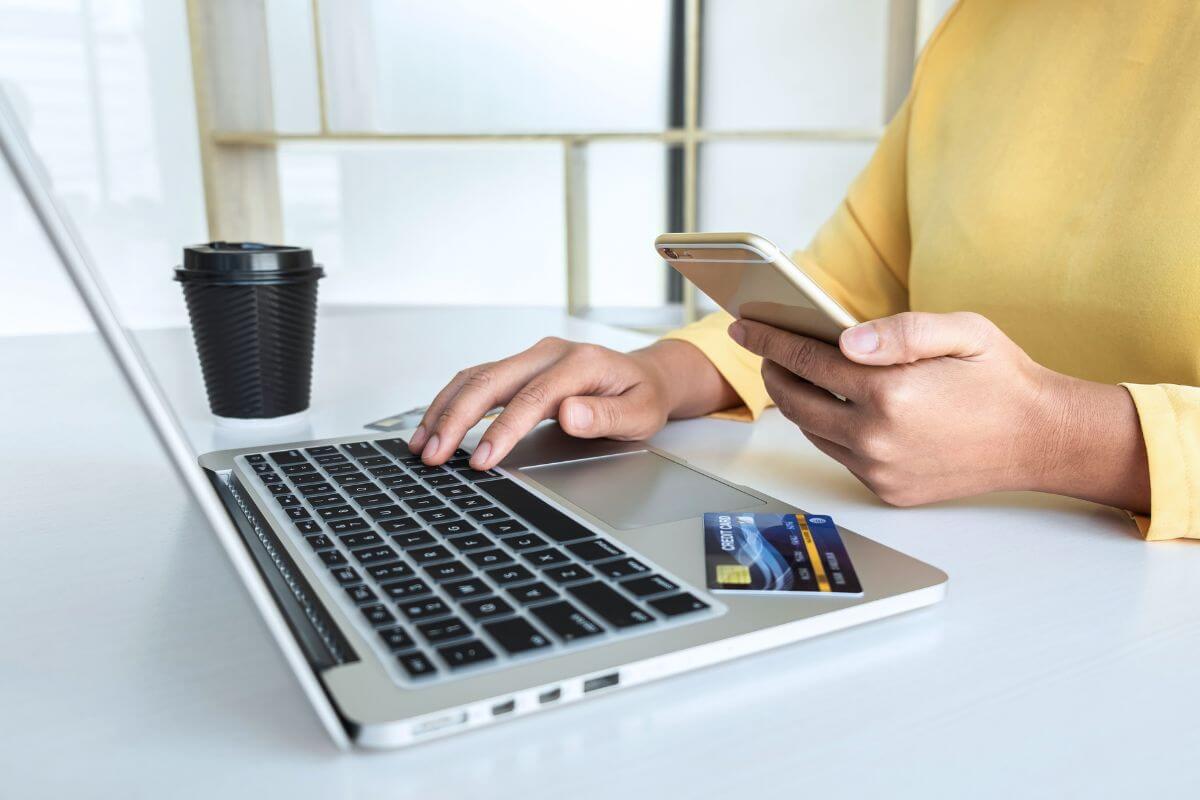 The photo tells about a coffee, a credit card, and a girl's hand holding a phone while using a laptop indicating to avoid credit card interest by paying balances in full to stay out of debt and save money.