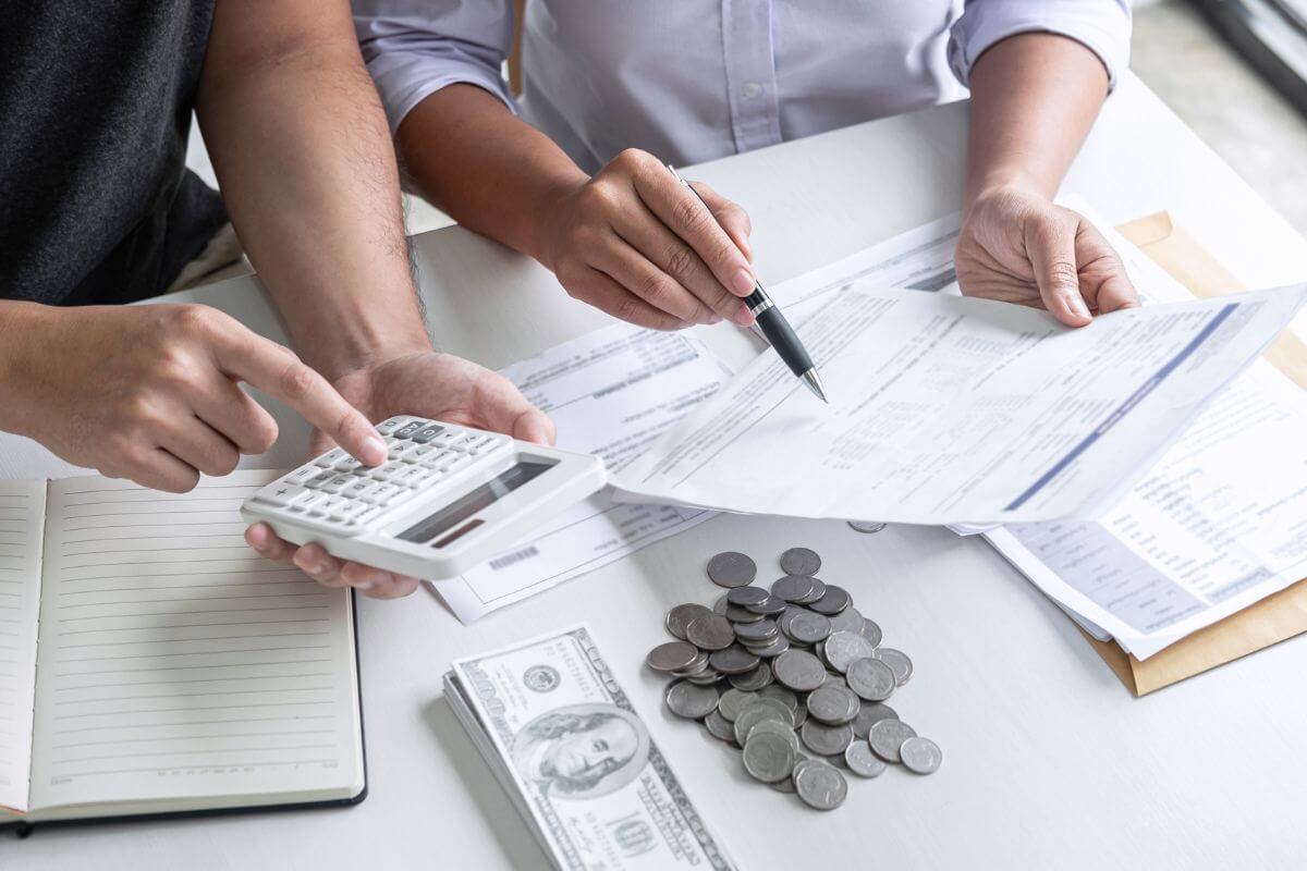 The image shows papers on the table, coins, dollar bills, a notebook, and a couple of calculating while referring to the paper to track and cut recurring expenses and use that money to boost your retirement contributions instead. 