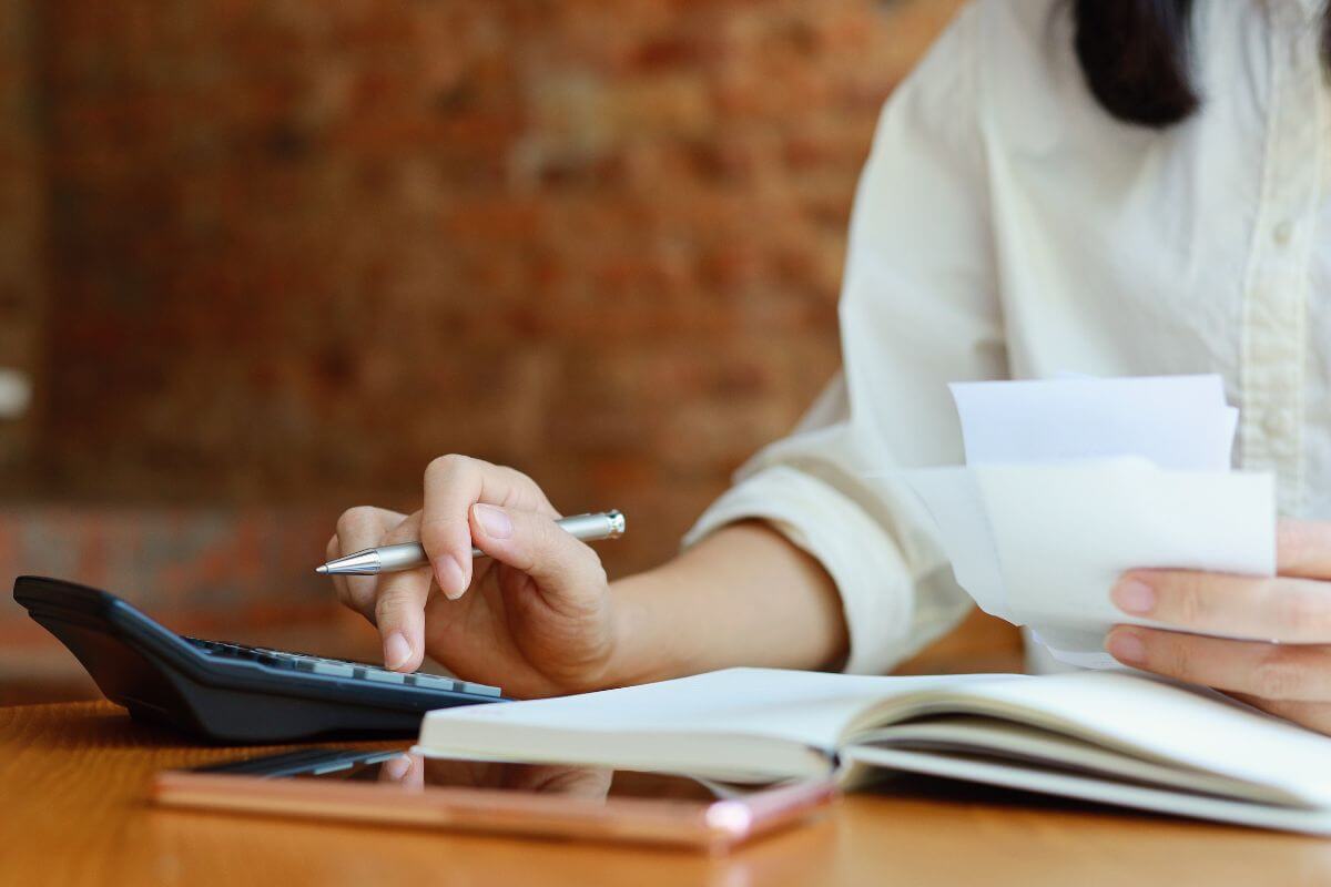 The image shows a phone, a notebook, and a girl's hand calculating while holding papers which means to create a detailed list of all debts helps you create solid repayment strategy to get out of debt faster.