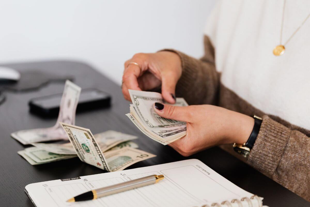 The photo tells about dollar bills on the table, a notebook, a pen, a phone, and a girl's hand counting dollar bills referring to reallocating funds from high-interest debt payments to retirement savings to build savings without adding to your budget.