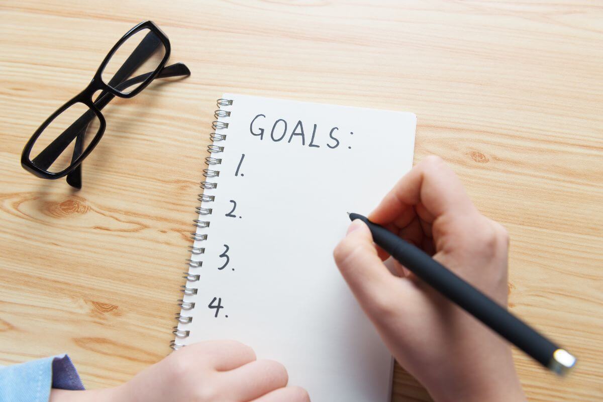 The photo tells about an eyeglass and a girl's hand holding a pen and writing in the notebook which means to set achievable goals that keep you motivated and focused as you work toward financial freedom.