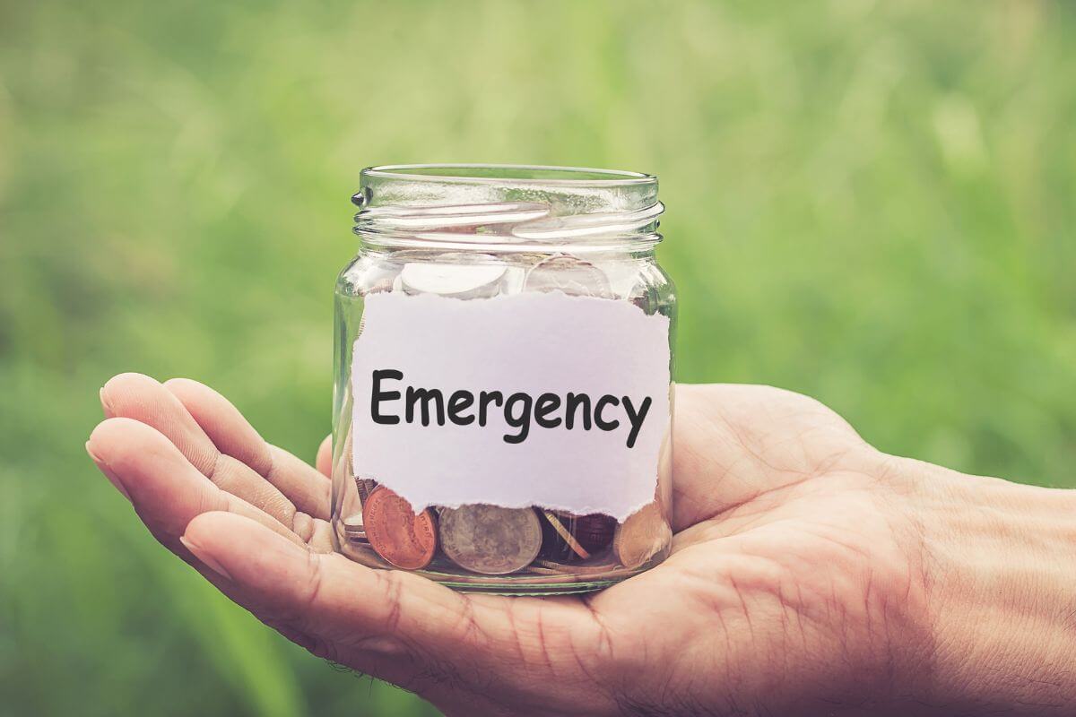 The picture tells about a man's hand holding a small jar filled with coins and with a note indicating to start by creating a small emergency fund that can help you cover unexpected costs without debt.