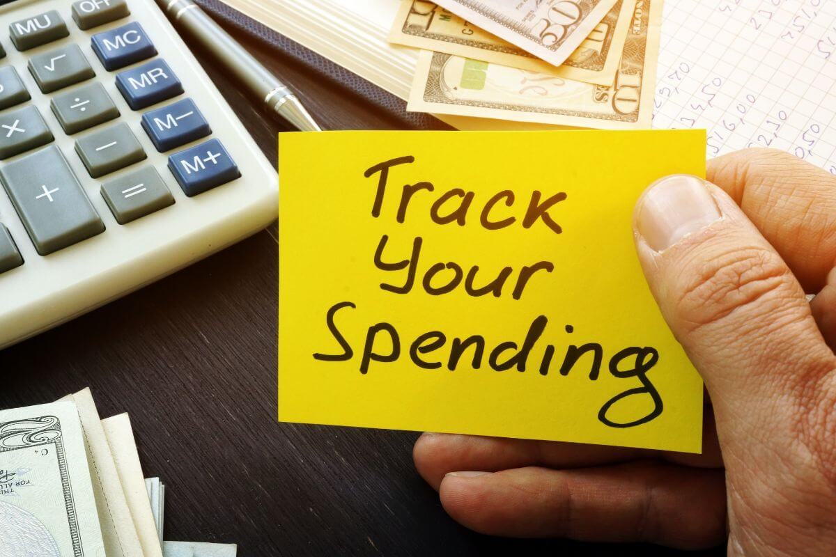 The photo tells about a calculator, a pen, dollar bills on the table, a notebook with numbers, and a man's hand holding a small piece of paper with notes indicating to track spending to find areas where you can cut back helps you see where your money goes.