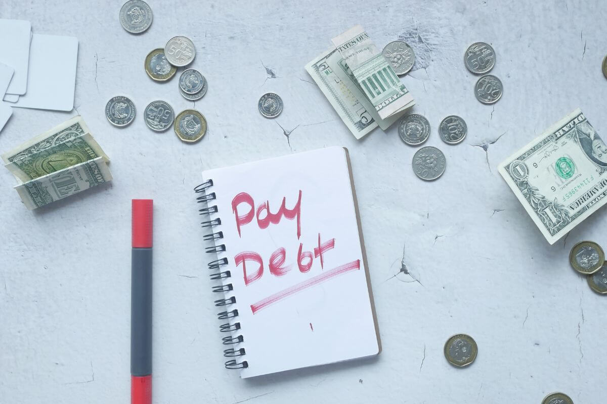 The image shows coins, dollar bills, a pen, and a notebook with notes referring to paying off high-interest debt to free up funds and build a stronger financial future.