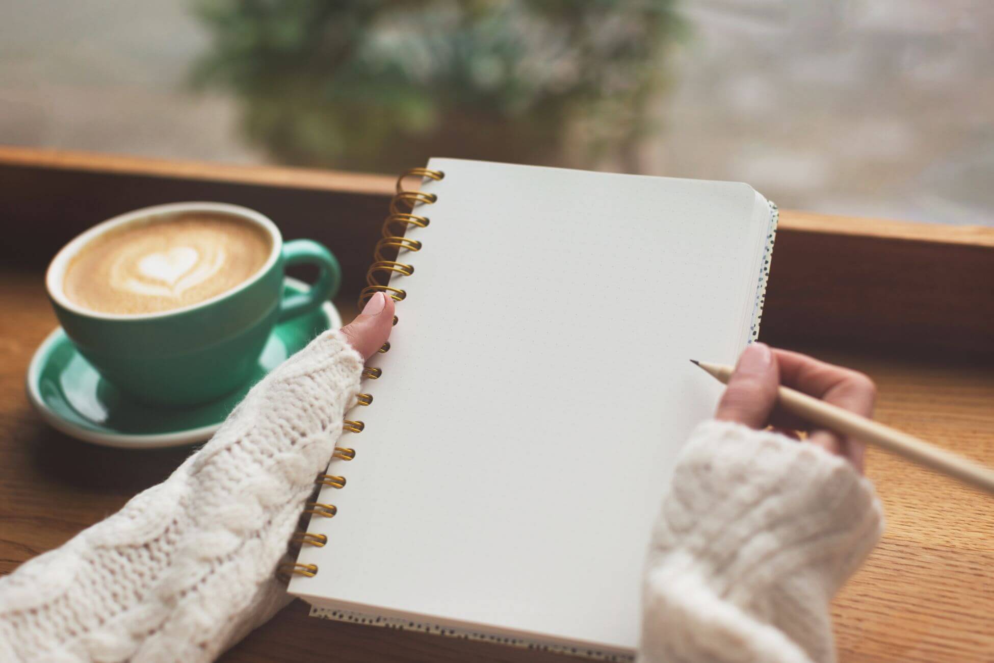 The image shows a coffee on the table and a girl's hand holding a pen and notebook which refers to setting realistic financial goals to build a strong safety net and helps you stay on track and build confidence.