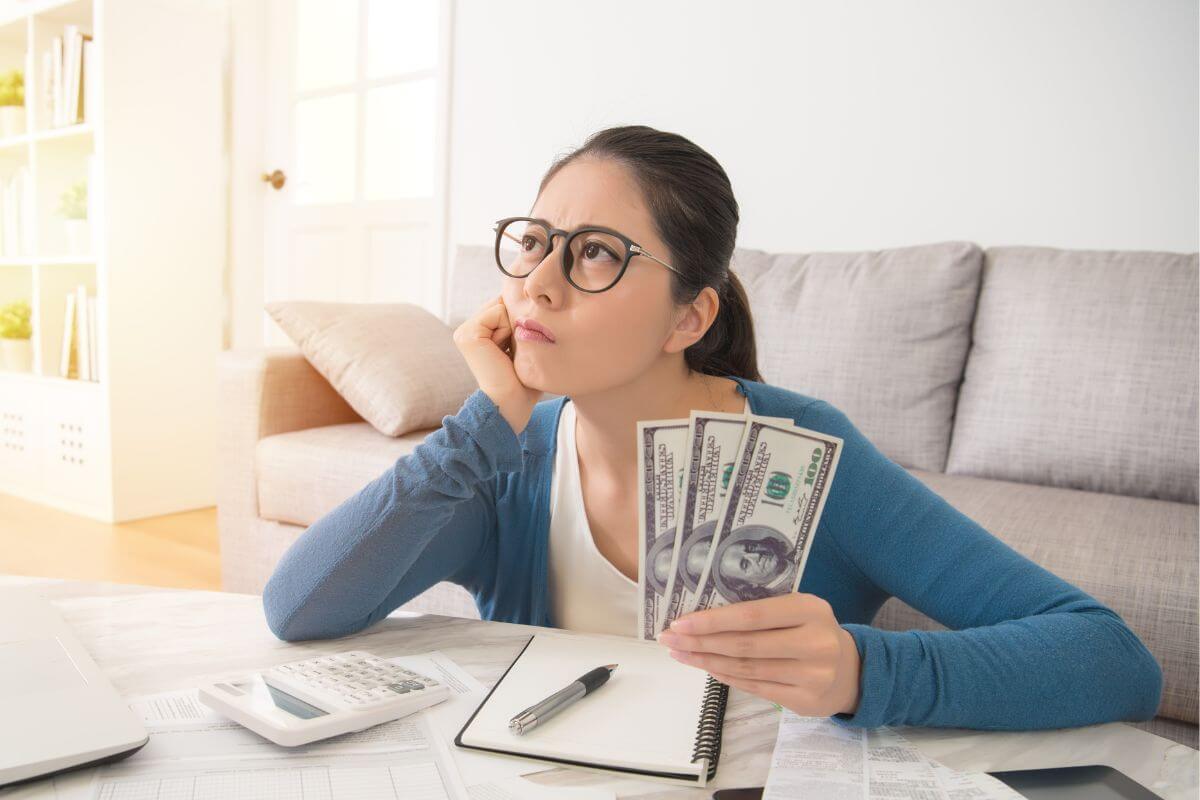 The photo tells about a calculator, a notebook, a pen, a laptop, and a girl holding dollar bills while thinking indicating that improving your money mindset can have a huge impact on your financial success.