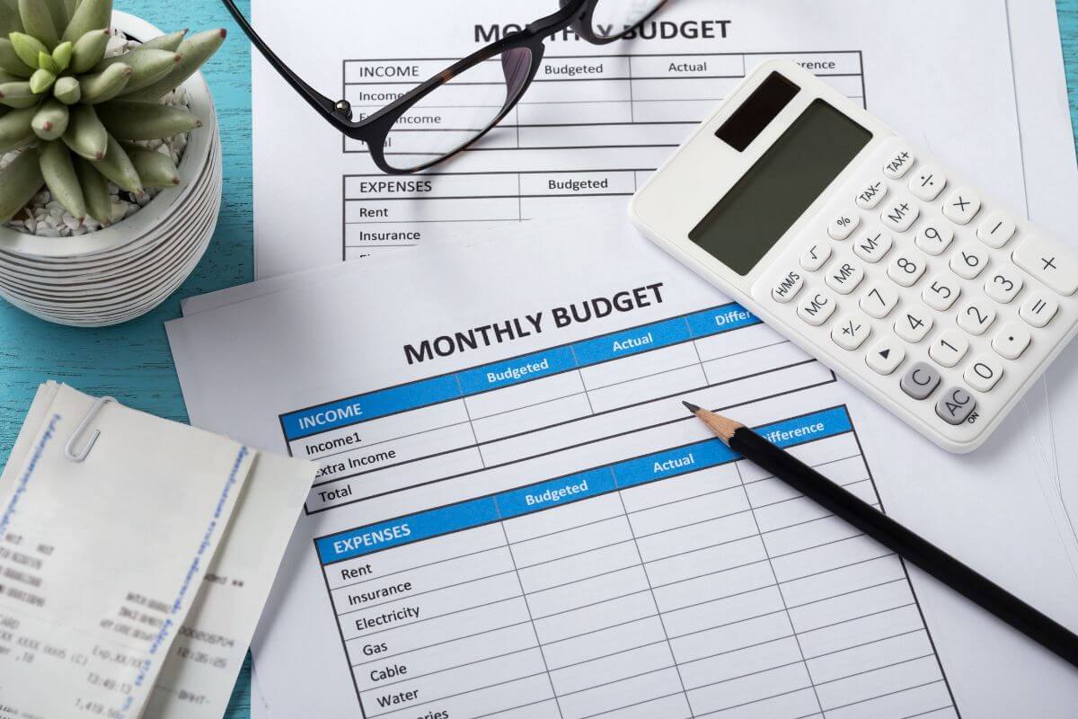 The picture tells about a calculator, a plant, an eyeglass, receipts, a pen, and papers with notes referring to creating a monthly budget to keep your spending clear and manageable.